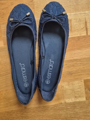 Mörkblå spetsballerinas från Esmara - Snygga mörkblå ballerinaskor från Esmara med spetsdetaljer och dekorativa rosetter framtill. Skorna har guldfärgade metalldetaljer på rosetterna och en klassisk rundad tå. Perfekta för dig som vill ha en feminin och stilren look. OBS. Lite stora i storleken så de passar bättre för sig med stl. 39.5-40.