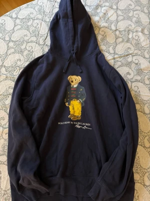Mörkblå hoodie Polo Ralph Lauren - Snygg mörkblå hoodie från Polo Ralph Lauren med Polo Bear-tryck på bröstet. Hoodien har dragsko i huvan och ribbade muddar. Tillverkad i mjuk bomullsblandning som känns skön mot huden. 