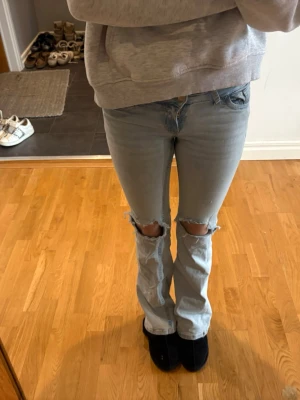 Trendiga Nelly Blå Jeans med Hål & Rosa Detalj - Säljer ett par ljusblå bootcut jeans med slitningar över knäna och broderade detaljer på bakfickorna. Jeansen har klassisk femficksdesign och är tillverkade i denim med en skön passform. Perfekta för en avslappnad och trendig look.