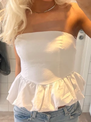 Vit volang tubtopp - Säljer en söt vit axelbandslös tubtopp med volangkant nedtill. Toppen har en croppad passform och är perfekt till jeans eller kjol. Tillverkad i ett mjukt bomullsmaterial som känns skönt mot huden. Enkel och stilren design som passar till många olika looks.