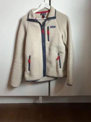 Schysst fleecejacka från Patagonia - Säljer en offwhite fleecejacka från Patagonia inmycket gott skicka, bara använt ett par gånger. Jackan har hög krage, två fickor fram och är tillverkad i mjuk teddyfleece. Perfekt för lager-på-lager och stiligt samt lätt att matcha med. En mer avancerad produktbeskrivningen ser du på sista bilden i annonsen. 