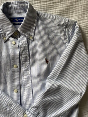 Randig skjorta från Ralph Lauren - Klassisk blå- och vitrandig skjorta från Ralph Lauren. Skjortan har knappar framtill, krage och långa ärmar. Perfekt för en stilren och tidlös look. Endast använd vid ett tillfälle, så nyskick. 