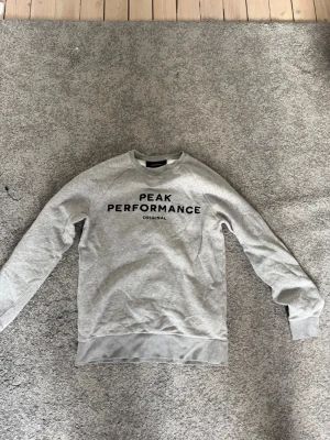 Grå sweatshirt Peak Performance M - Snygg ljusgrå sweatshirt från Peak Performance med svart logotyptext på bröstet. Klassisk rund halsringning, ribbade muddar vid ärmslut och nederkant. Perfekt för en avslappnad och sportig stil.