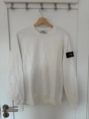 Vit sweatshirt från Stone Island - Vit sweatshirt från Stone Island med klassisk rund hals och ribbade muddar. Tröjan har den ikoniska svarta patchen med gul kompass på vänster ärm. Perfekt för en clean och stilren look.
