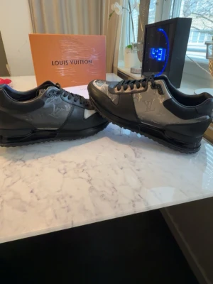 Louis Vuitton svarta sneakers - Snygga svarta Louis Vuitton sneakers i läder med gråa detaljer och präglad LV-logga på sidorna. Skorna har klassisk låg profil, svarta skosnören och en robust sula. Perfekta för dig som vill ha en exklusiv och trendig look. 