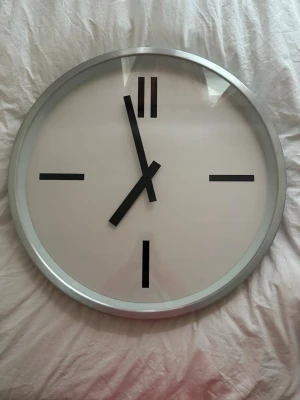 Stilren väggklocka i silver - Säljer en rund väggklocka med minimalistisk design. Klockan har en silverfärgad ram, vit urtavla och svarta markeringar för timmarna. Visarna är också svarta och tydliga. Perfekt för dig som gillar enkel och modern inredning.