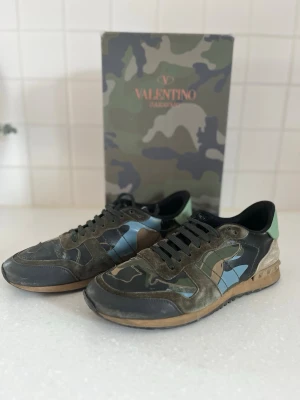 Valentino Garavani camo sneakers - Snygga sneakers från Valentino med camo-mönster i grönt, svart, beige och blått. Skorna har gröna skosnören, detaljer i mocka och läder samt en klassisk låg siluett. Perfekta för dig som vill sticka ut med exklusiv streetstyle.