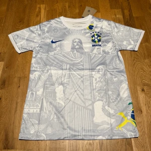 Brazil special jersey jesus  - Snygg vit fotbollströja från Nike med Brasilien-emblem och blå Nike-logga. Tröjan har unikt mönster med motiv från Brasilien, bland annat Kristusstatyn och tropiska detaljer. Rund halsringning och korta ärmar. Perfekt för dig som älskar fotboll och vill sticka ut.