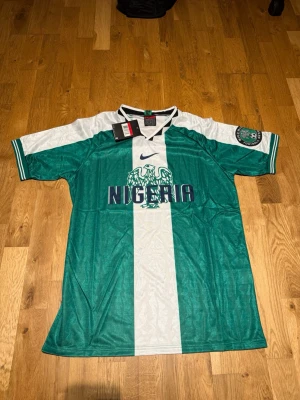 Nigeria home kit 1998 - Säljer en grön och vit Nigeria fotbollströja från Nike med klassisk retrodesign. Tröjan har ett stort örnemblem och texten 'Nigeria' på bröstet, samt klubbmärke på ärmen. Tillverkad i lätt och ventilerande material, perfekt för match eller träning.