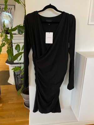 Svart klänning! Ny med prislappar!  - Stilren svart kortklänning med omlottdesign! Klänningen har draperade detaljer och ett mycket skönt material! Alma heter modellen! Storlek M/L