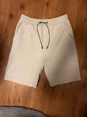 Beige shorts med snörning - Säljer ett par stilrena beige shorts med svart snörning i midjan och sidofickor. Perfekta för sommaren och har en relaxed passform som är både bekväm och enkel att matcha. Materialet känns mjukt och är troligtvis bomull eller polyester. Och en stor dragkedja ficka på höger bak 