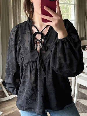 Svart blus med knytning - Svart blus i transparent tyg med mönster. Blusen har långa puffärmar och knytning framtill vid halsen. Passar även L!
