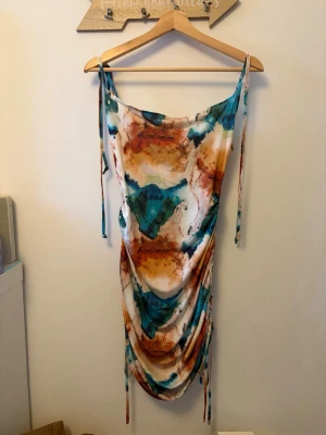 Färgglad kortklänning med snörning - Unik kortklänning med mönster i färgglada färger. Klänningen har smala axelband med knyt och snygg rynkning med snörning i sidorna. Materialet känns mjukt och stretchigt, perfekt för dig som vill sticka ut.