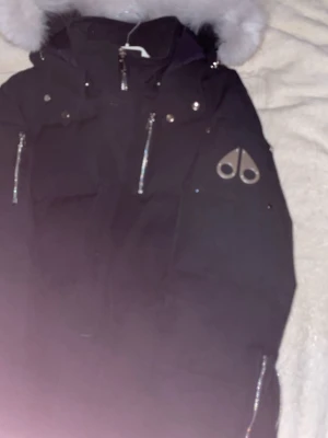 Svart dunjacka med päls från Moose Knuckles - Säljer en svart dunjacka från Moose Knuckles med silverfärgade detaljer och dragkedjor. Jackan har en stor huva med vit päls och det ikoniska Moose Knuckles-logotypen på ärmen. Perfekt för kalla vinterdagar och riktigt snygg design.