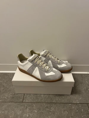 Maison margiela gats - Snygga sneakers i vitt och ljusgrått med ovandel i läder och mocka. Klassisk låg modell med gummisula och vita snören. Perfekta för dig som gillar stilrena och tidlösa skor med en retrovibe.