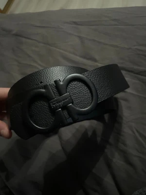  Ferragamo bälte - Säljer ett svart skärp från Salvatore Ferragamo med en bred, strukturerad läderrem och en ikonisk dubbelring-spänne i matt svart. Perfekt för att lyfta din outfit med en lyxig touch och cool design.