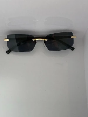 Svarta rektangulära solglasögon - Snygga solglasögon med rektangulära, svarta glas och tunna guldfärgade bågar. Glasögonen har minimalistisk design och svarta skalmar. Perfekta för dig som vill ha en stilren och modern look.