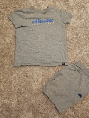 Grå shorts från Ellesse med snörning - Säljer ett par grå shorts från Ellesse med snörning i midjan och blå logga på vänster ben. Shortsen har ribbad midja och sidopaneler med blått tryck. Perfekta för en avslappnad stil.