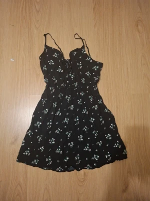 Svart blommig kortklänning - Svart kortklänning med små vita blommor och smala axelband. Klänningen har en lätt rynkad midja och ärmlös design, perfekt för varma dagar. Materialet känns som lätt bomull och ger en skön och luftig känsla.