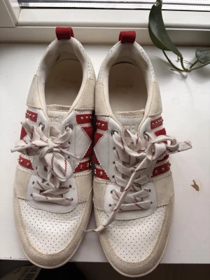 Vita och röda sneakers från Maje - Snygga sneakers från Maje i vitt och rött med stjärndetaljer och nitar. Skorna har perforerad tå, mockadetaljer och mönstrade skosnören. Klassisk låg modell med coola kontraster.