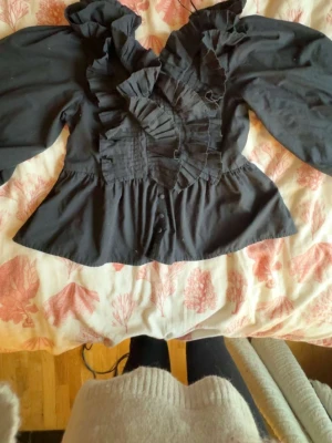 Svart blus med volanger och puffärm - Supersnygg svart blus med stora volanger framtill och puffiga långärmar. Blusen har knappar framtill och markerad midja för en feminin siluett. Perfekt för dig som gillar statement-plagg och trendiga detaljer. Materialet känns som bomull eller en bomullsblandning. Slutsåld💕