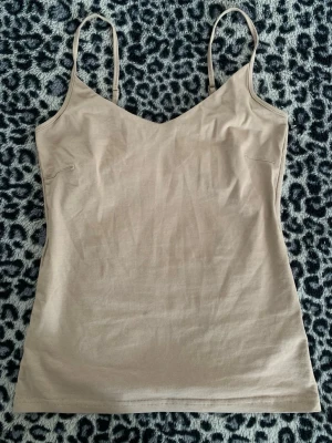 Beige linnetopp med smala axelband - Säljer en enkel och stilren beige linnetopp med smala axelband och v-ringad hals. Toppen är ärmlös och har en mjuk, lätt stretchig känsla som passar perfekt till sommaren. Perfekt att matcha med jeans eller kjol för en clean look.