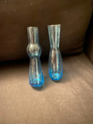 Två små blå glasvaser - Säljer två små vaser i blått glas med olika former. Den ena har en rundad midja medan den andra är mer avlång och smal. Perfekta för enstaka blommor eller som dekoration i hemmet. Snygg färg som ger ett fräscht intryck.