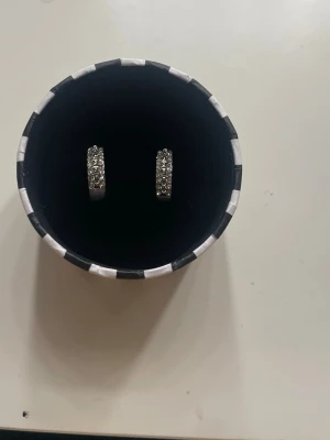 Silverfärgade hoops med stenar - Säljer ett par stilrena silverfärgade hoops med gnistrande klara stenar på framsidan. Örhängena har en klassisk rund form och stängs med klicklås. Perfekta för dig som gillar lite extra bling i vardagen.