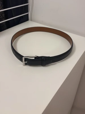 Svart skärp med silverfärgat spänne från Ralph lauren - Stilrent svart skärp med ett klassiskt silverfärgat spänne. Skärpet är slätt och har en brun insida, perfekt för att lyfta din outfit. Materialet är skinn och designen är enkel och tidlös. Från Ralph Lauren. Den är M i barn