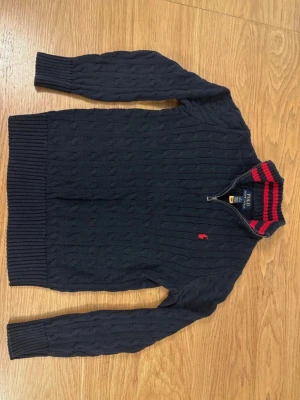 Mörkblå kabelstickad tröja Polo Ralph Lauren - Mörkblå kabelstickad tröja från Polo Ralph Lauren med halv dragkedja och ribbad krage med röda ränder. Tröjan har lång ärm, ribbade muddar och den klassiska röda logotypen broderad på bröstet. Perfekt för en snygg och avslappnad stil.