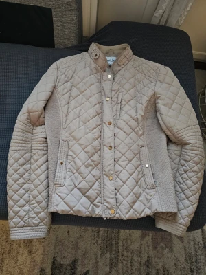 Beige quiltad jacka från Dea XL - Snygg beige quiltad jacka från Dea i storlek XL. Jackan har ståkrage med tryckknapp, guldfärgade detaljer, två framfickor och ribbade partier på sidorna och ärmarna för extra komfort. Perfekt för dig som gillar stilren och modern look.