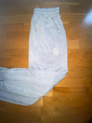 Grå mjukisbyxor från suvene  - Snygga grå mjukisbyxor från Suvene med broderad logga på benet. Byxorna har resår i midjan med dragsko och sidofickor. Tillverkade i mjukt bomullsmaterial som är perfekt för chill dagar. Klassisk passform och stilren look.