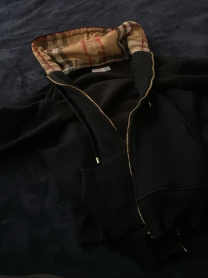 Svart hoodie från Burberry med mönstrad huva - Säljer en svart hoodie från Burberry med klassiskt rutmönster på insidan av huvan. Hoodien har dragkedja, fickor framtill och snören vid huvan. Materialet är mjukt och bekvämt, perfekt för en chill look. Märket syns tydligt på etiketten.