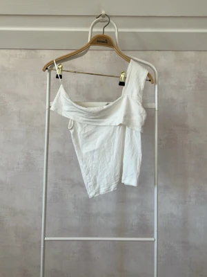 Vit oneshoulder topp - Säljer en vit oneshoulder topp med asymmetrisk skärning. Toppen är ärmlös på ena sidan och har bred axelrem på andra sidan. Perfekt för dig som vill ha en stilren och trendig look.