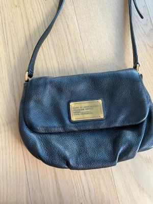 Svart axelväska från Marc by Marc Jacobs - Snygg svart axelväska i läder från Marc by Marc Jacobs med justerbar axelrem och guldfärgad metallplatta framtill. Väskan har en mjuk, rundad form och diskret struktur i materialet. Perfekt för dig som gillar klassiska designer och stilrena detaljer. Endast använd två gången och i mycket bra skick utan direkta tecken på användning. 