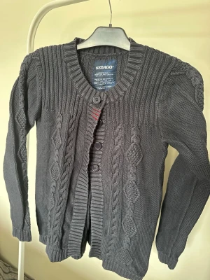 Mörkgrå kabelstickad kofta Sebago - Mörkgrå kabelstickad kofta från Sebago med ribbade detaljer vid axlar, krage och muddar. Koftan har knappar framtill och ett diskret broderi på ryggen. Långärmad och tillverkad i mjukt material som passar perfekt för lager-på-lager.