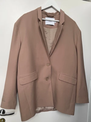 Oversize kavaj 34-38 - Snygg och trendig oversized kavaj i beige/ cognacbrun  nyans från Junkyard. Modellen är enkelknäppt med två knappar framtill och har klassiska fickor med lock. Kavajen är dessutom snyggt fodrad, vilket ger en extra kvalitetskänsla och en fin finish på insidan. Perfekt både till vardag och för en mer uppklädd outfit – lika fin till jeans som över en klänning. Det är en mycket oversized XS/S så den  passar även M/L Mått :  Längd 80 cm  Bröst och midja 60 cm ( platt mätt)  Armlängd från axel 62 cm 