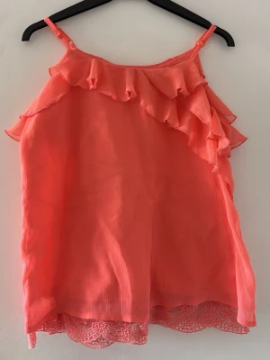 Korallrosa linnetopp med volang - Säljer en snygg korallrosa linnetopp från LC Waikiki med volangdetaljer över axeln och fram. Toppen har tunna axelband och ett skirt, lätt material med spetskant nedtill. Perfekt för sommarens varma dagar.