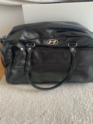 Svart weekendbag från Hyundai - Snygg svart weekendbag i glansigt konstläder med Hyundai-logga framtill. Väskan har dubbla handtag, dragkedja upptill och en stor ytterficka med dragkedja. Perfekt storlek för övernattning eller gymmet och har en stilren, enkel design.