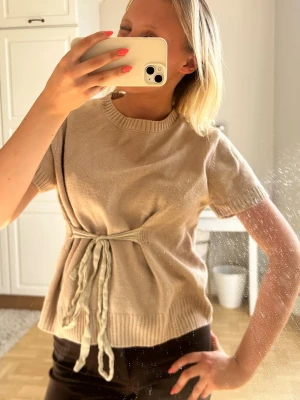 Beige stickad topp med knytband💖💖 - Trendig beige stickad topp med korta ärmar och ribbad halsringning. Toppen har ett dekorativt knytband i midjan som ger en snygg siluett. Perfekt att styla med både jeans och kjol för en avslappnad look.