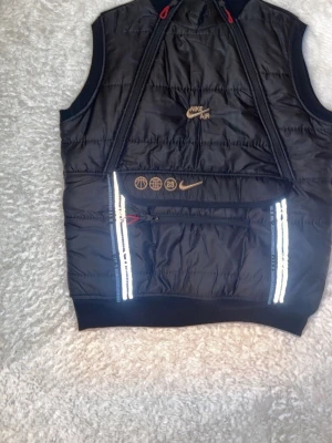 Svart Nike Air dunväst med reflexdetaljer - Snygg svart dunväst från Nike Air med broderade loggor och symboler i guld, reflexdetaljer vid dragkedjorna och praktiska fickor. Västen har sportig look och är perfekt för lager-på-lager-stil. Materialet är glansigt och lätt vadderat.