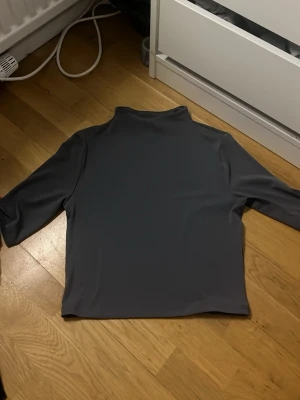 blå grå polotopp med trekvartsärm - Säljer en stilren mörkgrå polotopp med trekvartsärm. Toppen har en hög krage och är gjord i ett slätt syntetmaterial som känns mjukt mot huden. Perfekt för dig som gillar minimalistisk och clean look.
