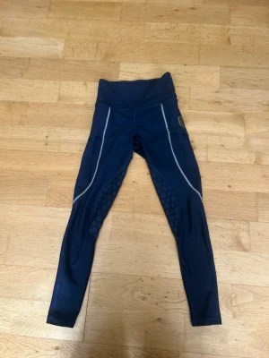 Mörkblå ridtights från Maya delorez - Snygga mörkblå ridtights från Maya delorez med hög midja och silikongrepp på insidan av benen. Tightsen har vita detaljer längs sidorna och logotyp på höften. Perfekta för träning och ridning, tillverkade i stretchigt material som ger bra rörelsefrihet.