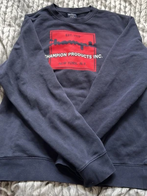 Svart sweatshirt från Champion - Cool svart sweatshirt från Champion med stort rött och vitt tryck på bröstet där det står 'Champion Products Inc. New York N.Y.' och 'Est. 1919'. Klassisk rund halsringning och långa ärmar. Perfekt för en avslappnad streetwear-look.