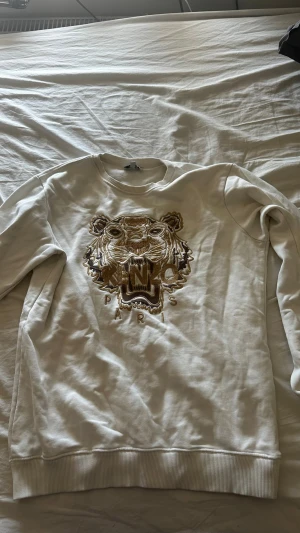 Vit Kenzo hoodie med tigerbroderi - Säljer en vit hoodie från Kenzo med stort broderat tigerhuvud och texten 'KENZO PARIS' på bröstet. Tröjan har ribbade muddar och är gjord i mjukt bomullsmaterial. Perfekt för dig som vill ha en unik och trendig look.köpt ny för 2400kr