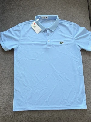 Ljusblå polotopp från Lacoste M - Säljer en ljusblå polotopp från Lacoste med klassisk krage och knappar framtill. Toppens ikoniska krokodillogo är broderad på bröstet. Materialet är bomull och passformen är normal. Perfekt för dig som gillar stilrena och sportiga plagg.