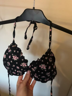 Svart blommig bikini med volang - Söt svart bikini med små rosa blommor och volangkant. Överdelen har knyt i nacken och ryggen för justerbar passform. Underdelen har matchande volangdetaljer. Perfekt för sommaren och strandhäng! ENDAST TESTAD!