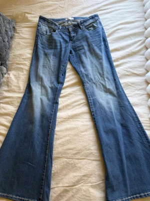 Blå bootcut jeans - Säljer ett par blå bootcut jeans från i storlek 40. Jeansen har låg midja och snygg utsvängd passform. Materialet är mjukt denim med ljusa slitningar framtill för en cool vintagekänsla.