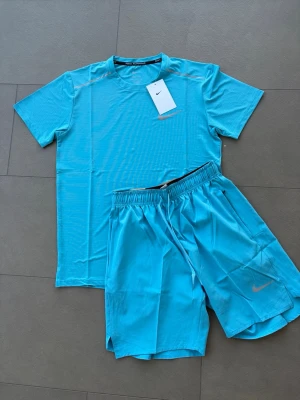 Nike Dri-FIT träningsset turkos - Nike Dri-FIT träningsset i turkos, perfekt för löpning eller gym. Setet består av en kortärmad t-shirt och matchande shorts med elastisk midja. Båda plaggen har Nike-logga och är tillverkade i ett lätt, snabbtorkande funktionsmaterial.