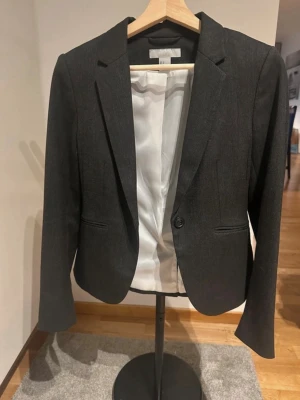 Mörkgrå kavaj med klassisk passform - Säljer en mörkgrå kavaj från H&M med klassisk passform och ett stilrent snitt. Kavajen har ett knappstängning framtill, två fickor och ljus innerfoder. Perfekt för dig som vill ha en enkel och snygg look.